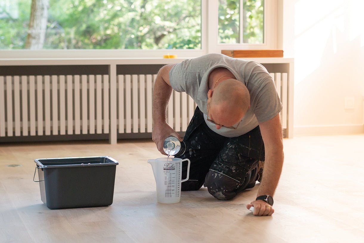 Person pouring Rubio Monocoat wood cleaner in a measuring cup on a natural wood floor.