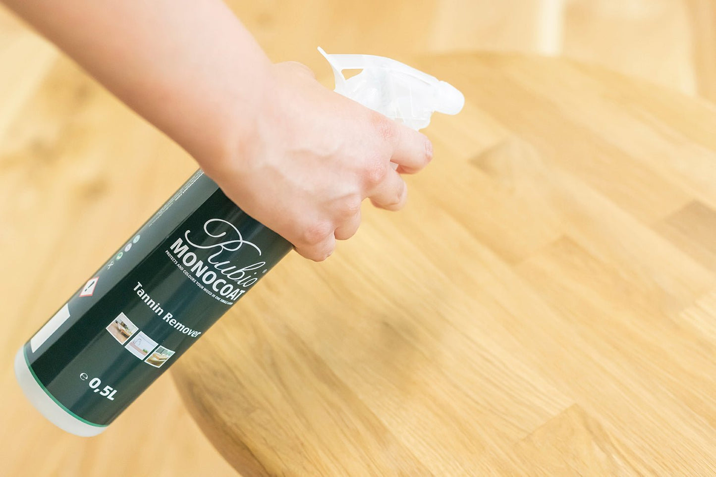 Hand holding a spray bottle labeled 'Rubio Monocoat' on a wooden floor.